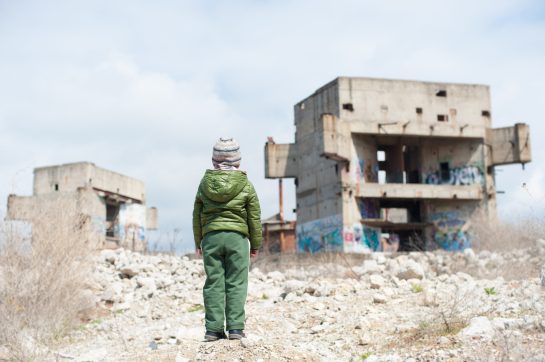 one little child in green jacket standing on ruins of destroyed buildings in war zone