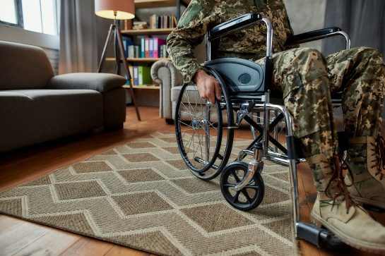 Cropped shot of disabled military man wearing camouflage in a wheelchair during therapy session in the living room. PTSD concept. Horizontal shot. Focus on hand and wheelchair